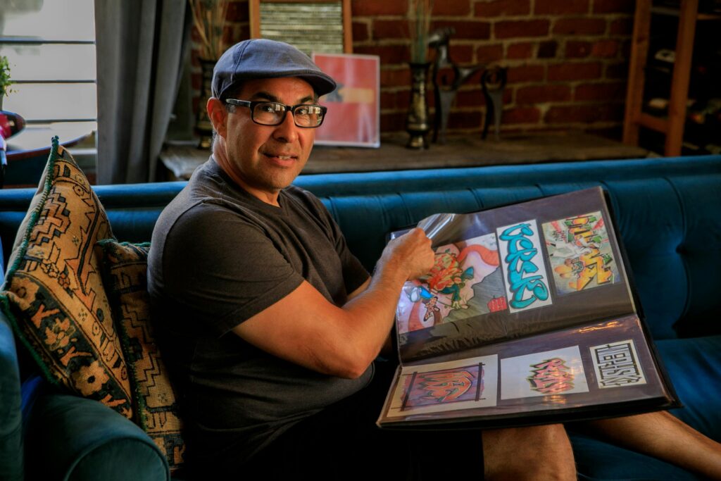 Man in hat and glasses proudly displaying his colorful graffiti art portfolio on a couch.